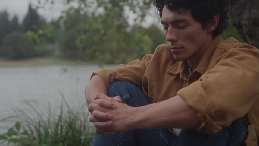 A man with dark curly hair sitting on the ground with his knees drawn up arms clasped over his knees and his eyes closed or looking down in a posture of reflection against a backdrop of water and dark trees