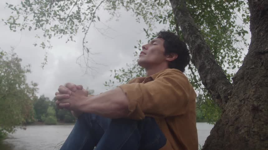The video is a lowangle outdoor shot of a man sitting under a tree by a river or pond on an overcast dayhe is resting with his eyes closed and hands clasped appearing to be in a state of meditation or peaceful reflection surrounded by nature