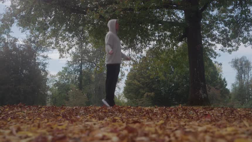 The video shows a lowangle wide shot of a man in a white hooded sweatshirt and dark pants shadowboxing or skipping rope without a ropehe is exercising outdoors in a park or forest clearing surrounded by green trees and a carpet of reddish