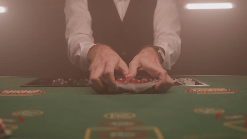 The hands and forearms of a dealer wearing a white shirt and dark vest as he perform a riffle shuffle with two stacks of playing cards on a green felt gaming table in a casino setting