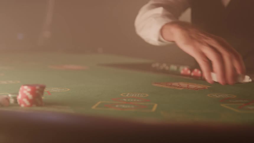 The video is a lowangle mediumcloseup shot of a dealers hands on a green felt poker or blackjack tablethe dealer wearing a white shirt and dark vest collects chips and cards in a smoky dramatic casino environment