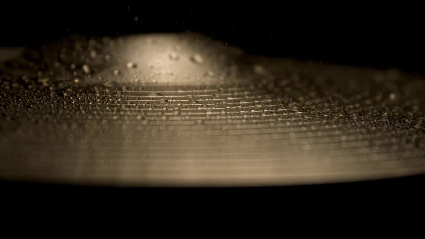 A close dramatic view shows a wet goldtoned cymbal vibrating and shaking off large water droplets against a dark black background
