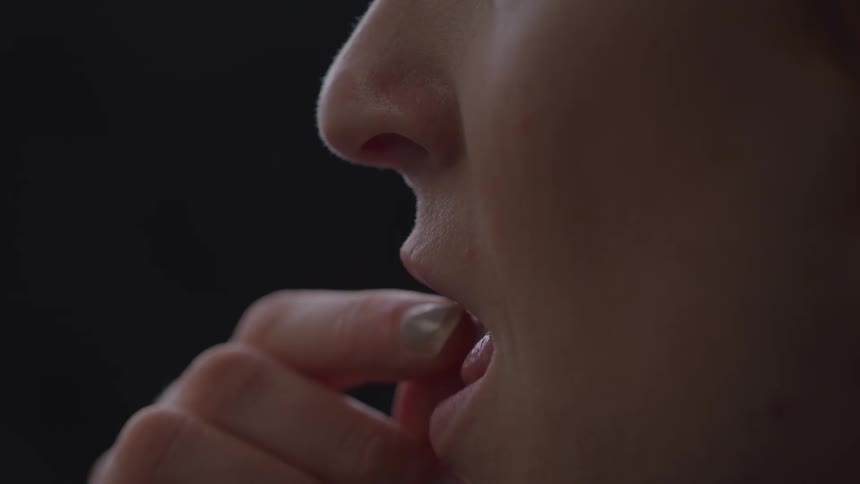 A close profile view shows a persons mouth and nose as they take a pill or capsule with a manicured hand against a dark background