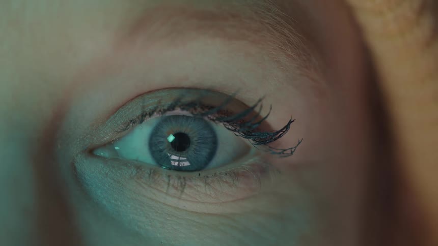 A camera is focused extremely close on a persons open eyeball which is facing forward and reflecting light sources on its surface while the surrounding skin remains motionless