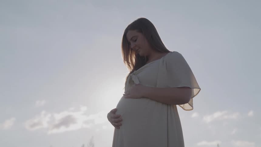 Smiling expectant woman briefly obscured by the glaring sun hugs her abdomen