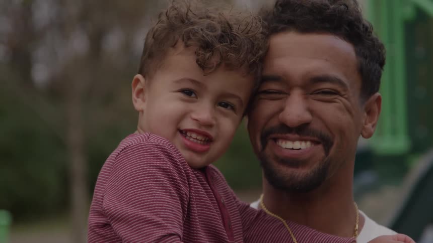 An adult man holding a young boy with both the man and the child positioned close together looking directly at the camera and smiling broadly in an outdoor setting