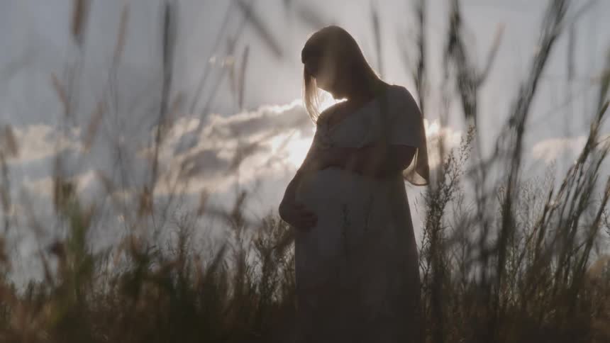 A pregnant woman rubs her abdomen while standing in a grassland on a partly sunny day