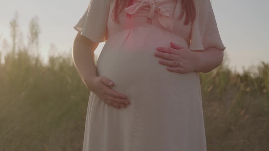 The hands and belly of a pregnant person wearing a lightcolored dress as she gently rest both hands on the abdomen standing in an outdoor field bathed in soft backlighting