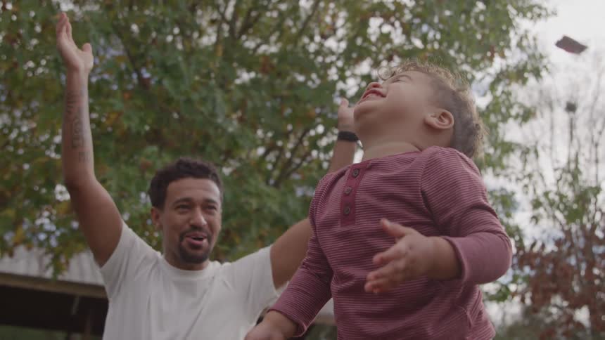 Father smiles while watching his son throw fallen leaves into the air in the yard of their house on an autumn day