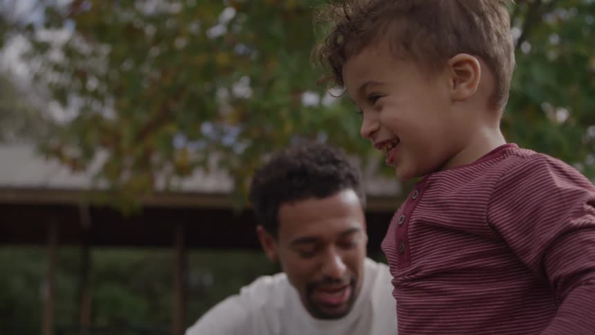A joyful toddler throws autumn leaves in the air while his father watches with a smile