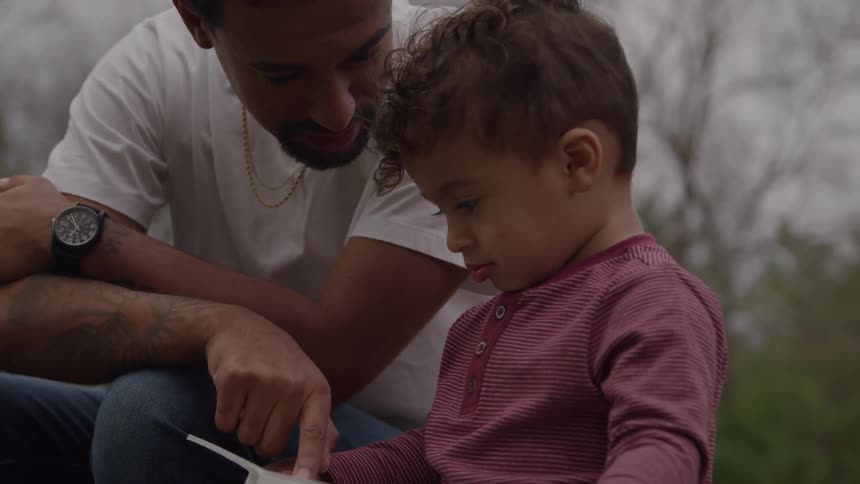 An adult man and a young child sitting closely together outdoors with the man leaning over the child holding a small book and pointing to the pages while the child looks down at the book