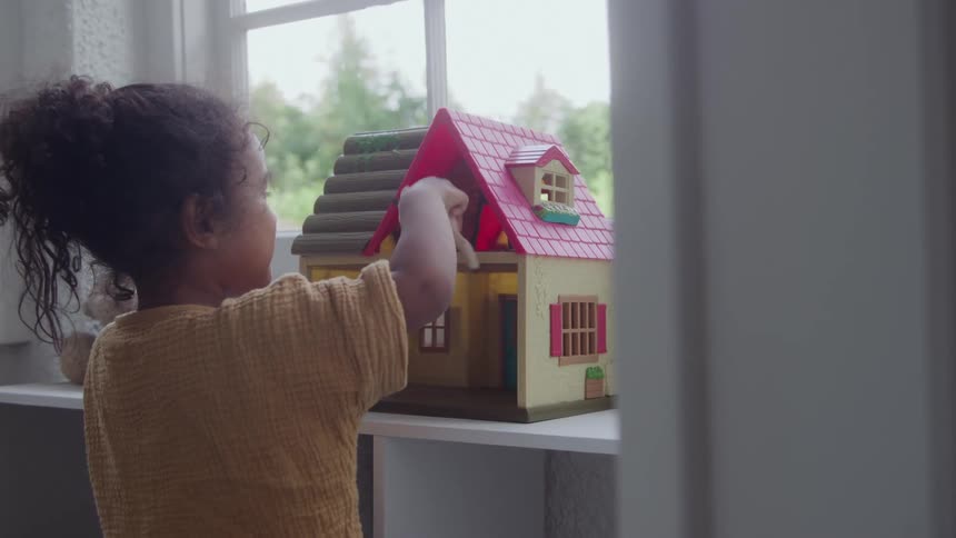 A young girl plays with a dollhouse placed on a shelf beside the window at midday