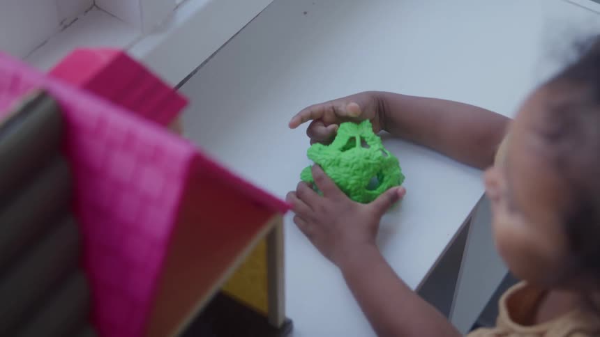 Girl plays with two green toys near a bookcase holding a dollhouse