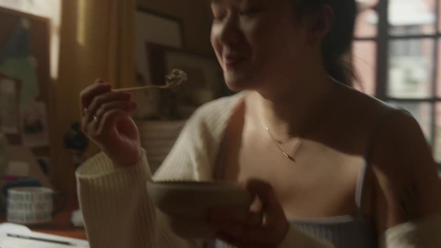 A young woman sits at a desk in a room with a window holds a ceramic bowl in one hand and uses a spoon with the other hand to scoop food from the bowl and eat