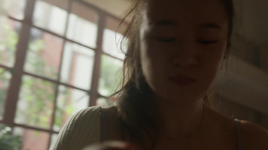 A young woman holds a piece of tableware in their hand and raises it to their face to ingest food from a bowl while dancing near a window with sunlight streaming into the room