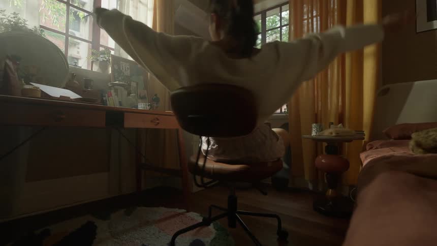 A young woman sits in a rolling office chair in an indoor room with two windows and extends both upper limbs directly upward above the head then lowers the arms and she has both feet on the chairs base