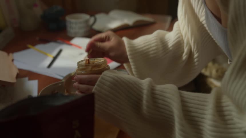 A teenage girl wearing a light sweater holds a mason jar with one hand and inserts a small spoon full of spread in a food bowl in a bedroom setting