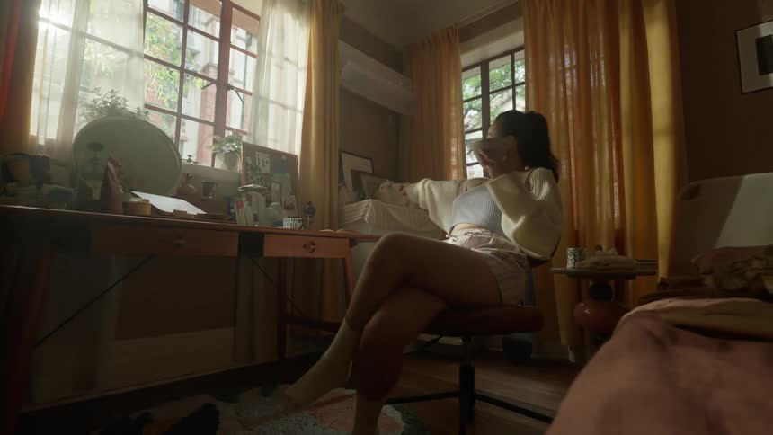 A teenage girl is seated in an adjustable chair at a wooden table in an indoor room holding a container near their mouth and spinning the chair while facing two curtained windows