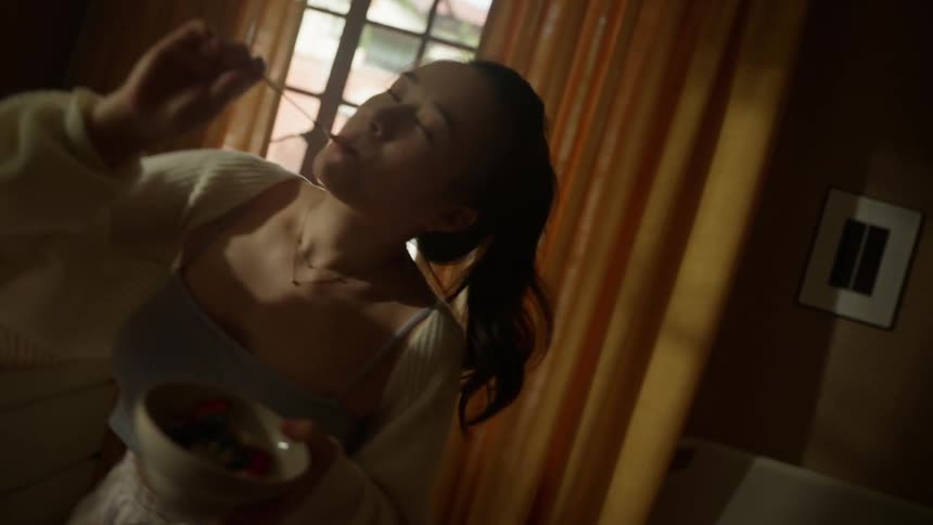 A young woman stands in an indoor space with draped curtains and windows uses a spoon to grab food from a small bowl and moves the upper body in a slow dance while chewing