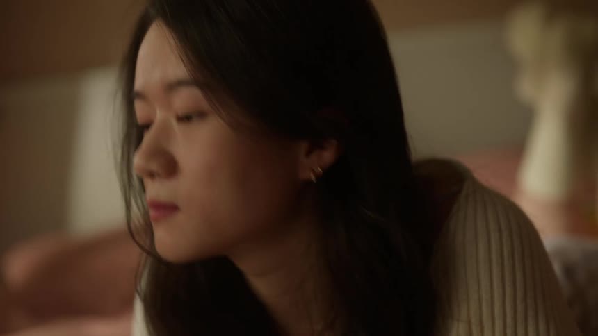 A young woman lying on the bed wearing sweater and socks turn around smiles and grab something off screen in a bedroom setting