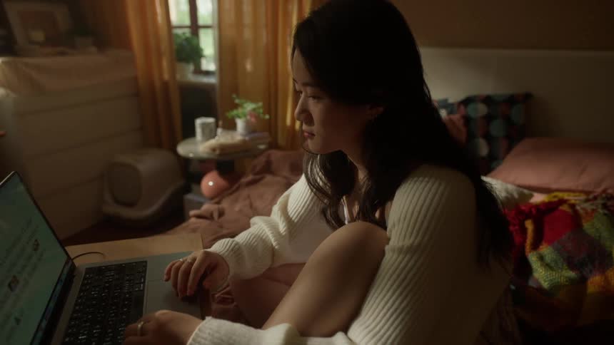 A young woman is sitting on a bed with a small piece of furniture holding a computing device operating the device with both hands before closing the lid and reclining onto the bed