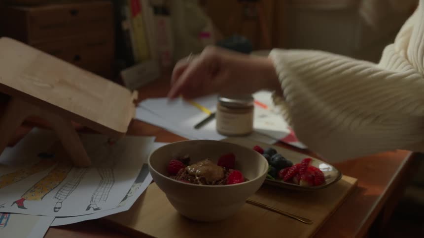 A woman with dark hair tied back wears a cream colored sweater and stands at a cluttered wooden desk to place berries on a breakfast bowl next to drawings and a small wooden easel in a sunlit room