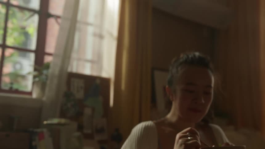 A teenage woman is dancing indoors near a window then sits and uses a spoon to eat something from a bowl in a bedroom setting