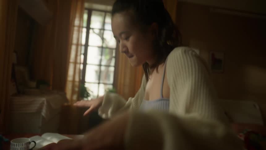 A teenage girl dances in front of a messy desk in a bedroom setting with windows and furniture