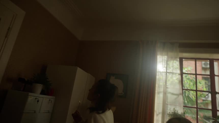 A young woman holding a small bowl is positioned between two windows and moves her body rhythmically in a dimly lit indoor space with objects and furniture surrounding her