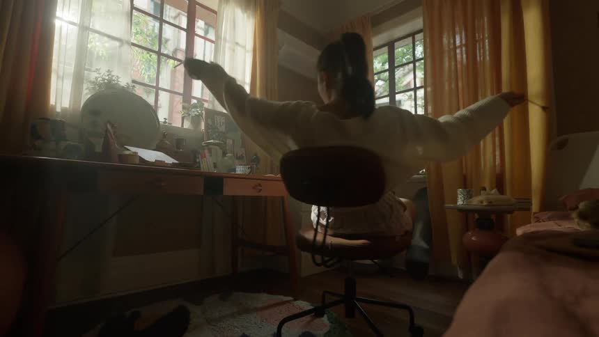 A teenage girl sits on a rolling chair with her feet crossed on the seat eats from a small round container then stretches her arms and continues to sit while holding the container