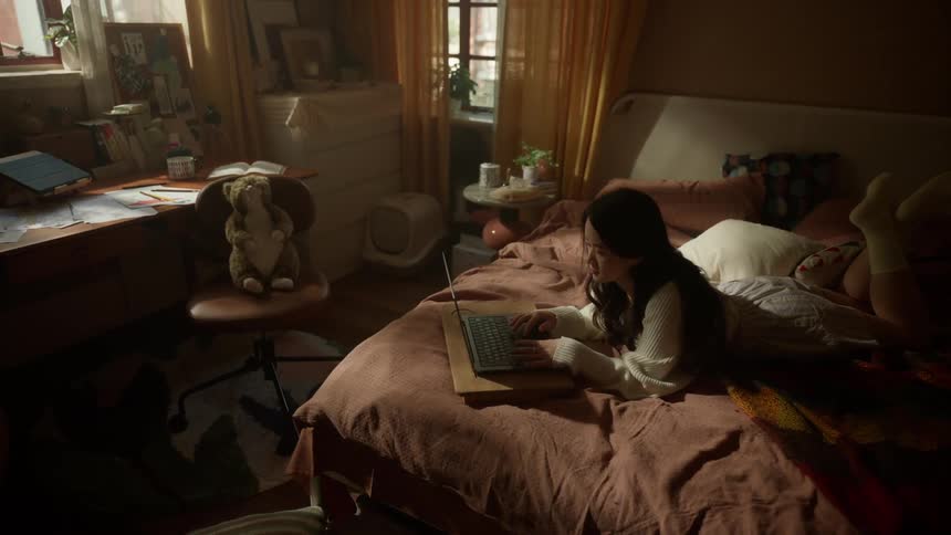 A young woman lies on her bed at midday focused on her laptop as sunlight streams through the bedroom window casting soft light across the room