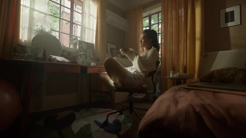 A woman wearing a white knit sweater and shorts sits cross legged in a desk chair stretching her arms and enjoying a drink in a warm sunlit bedroom with a desk by the window