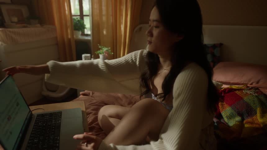A teenage girl sits on a bed using a laptop on a small tray then pushes the computer away stretches and go out of the frame