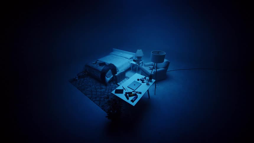 An elevated wide view shows a brightly lit bed a desk with a laptop and a bedside chair and lamp on a rug all isolated in a vast uniformly dark blue and shadowy room