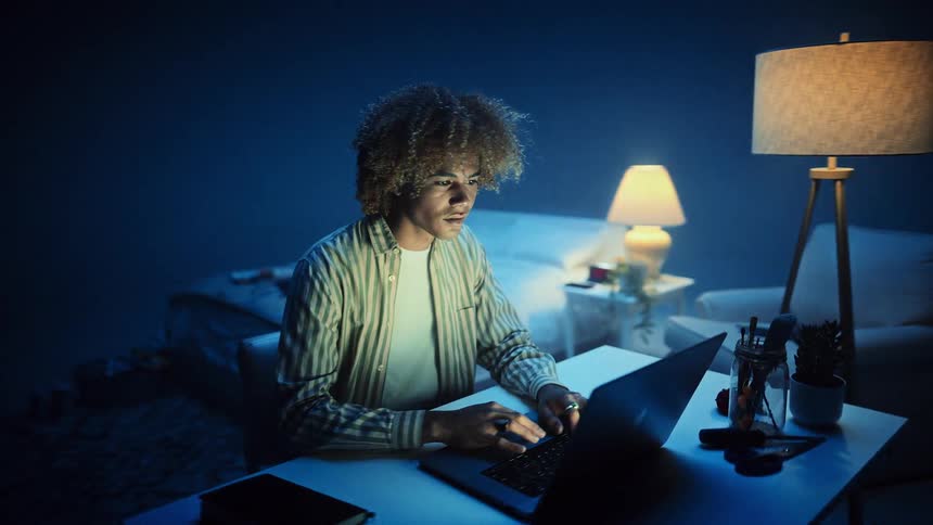 Person sitting at a desk using a laptop in a room lit by blue light and a lamp