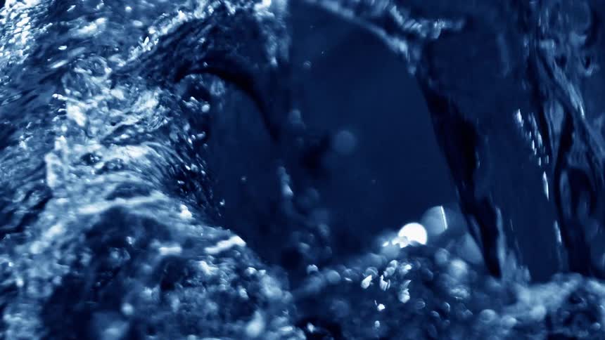 A closeup view shows a turbulent blue liquid as it flows and splashes with many bright white highlights reflecting off the surface