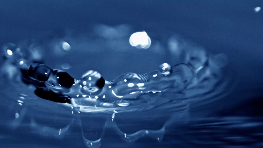 A crown shaped splash rises from dark blue water as a droplet hits the surface