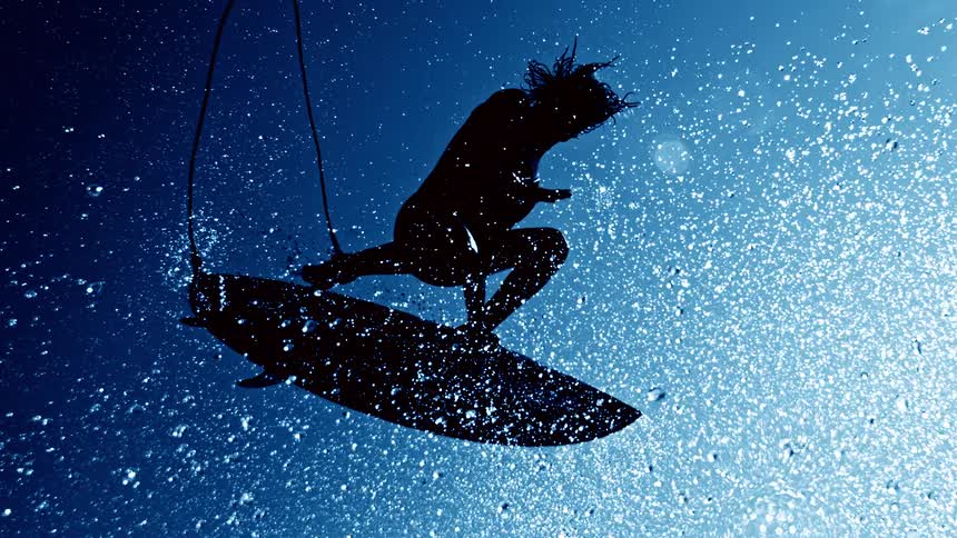 A striking silhouette shot from below shows a male surfer suspended in the air on a surfboard against a deep blue sky surrounded by brightly lit water droplets and spray