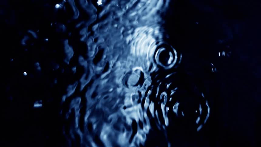A series of concentric ripples shimmer across dark blue water as droplets fall and disturb the surface
