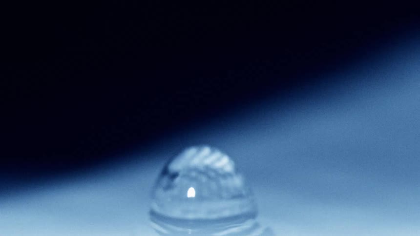 A water droplet falls onto still blue water creating ripples in a dark atmosphere