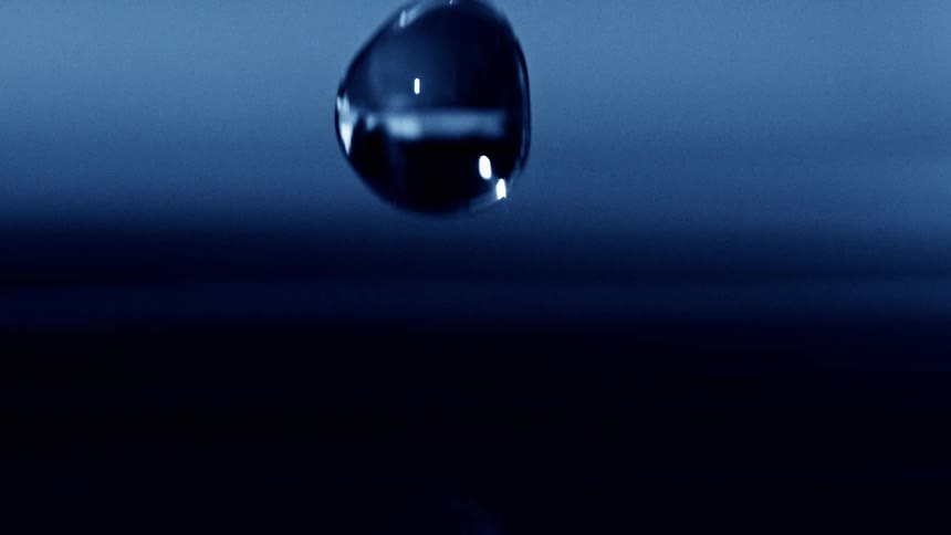 A water droplet falls and splashes on a glossy dark surface with its reflection glowing in cool blue light