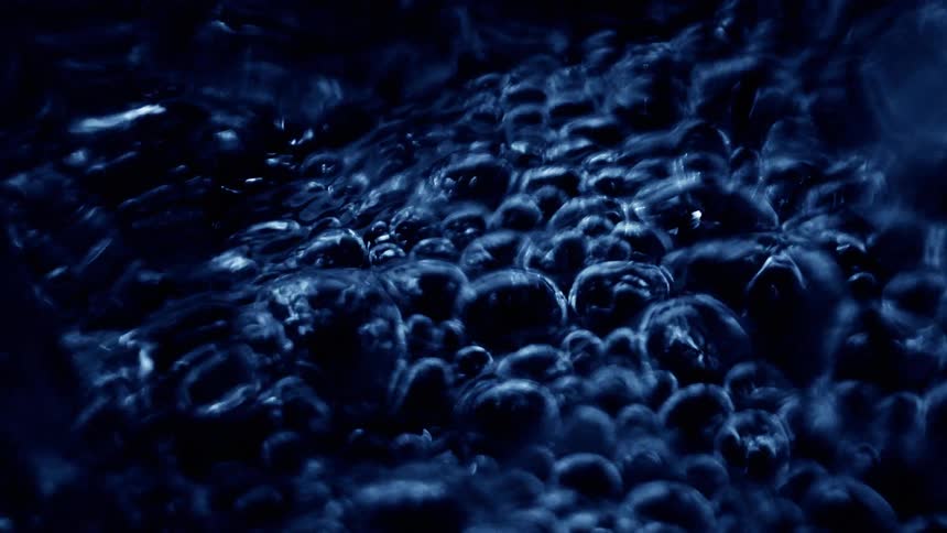 A series of bubbles form in a liquid spread across a dark surface rising and clustering with subtle shimmer