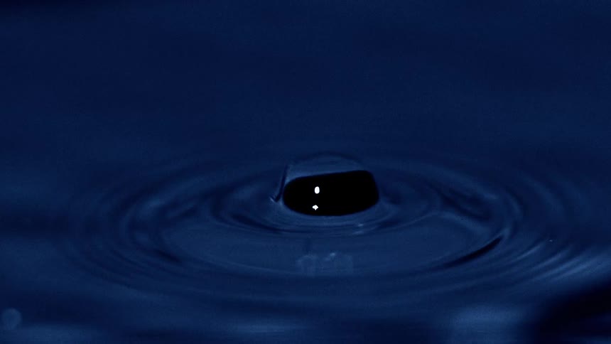 A water droplet falls onto still water triggering a ripple effect that gently expands across the surface