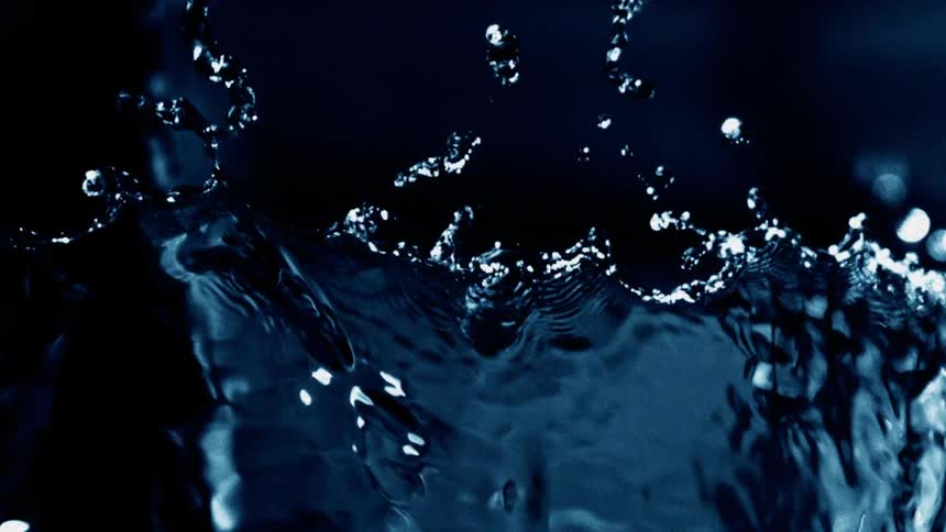 A high speed close up captures dark blue water flinging upward in curved tendrils against a soft glowing spot of light