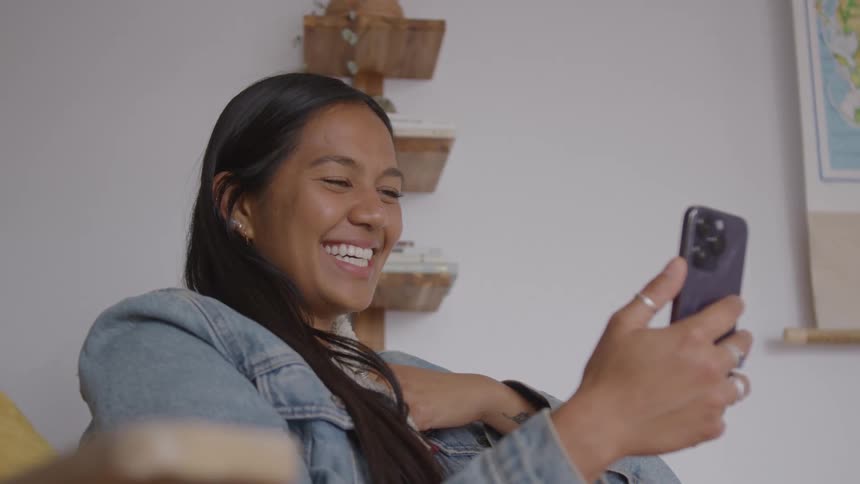 A female smiles widely as she looks at her phone facing her as she sits in a room