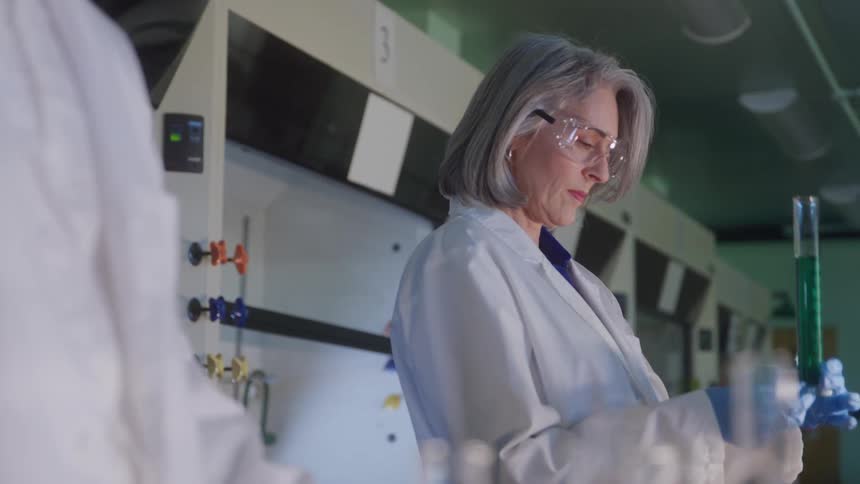 A focused woman scientist wearing safety goggles examines a test tube of green liquid in a laboratory