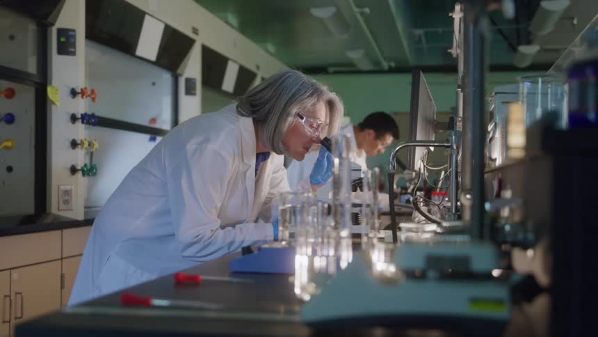 The video shows two scientists working in a laboratory both wearing lab coats and safety glassesa woman in the foreground is leaning over a lab bench examining glassware near a fume hood while a man is visible in the background looking a