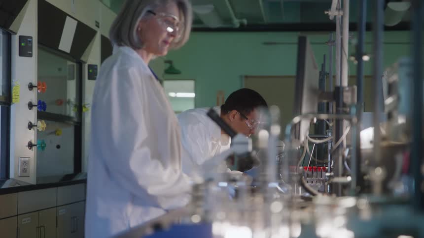 A man in safety goggles works focused at a lab bench while a woman in a white coat stands in the foreground