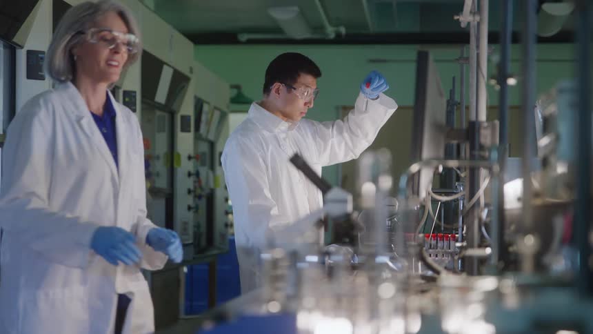 In a science lab a male researcher holds a test tube up to the light for examination while his female colleague walks into the foreground