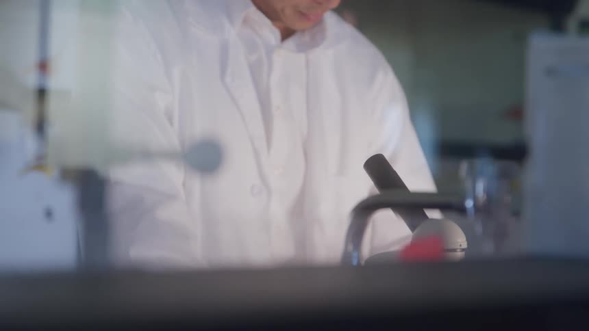 A man in safety goggles peers into a microscope with concentrated attention at a lab bench